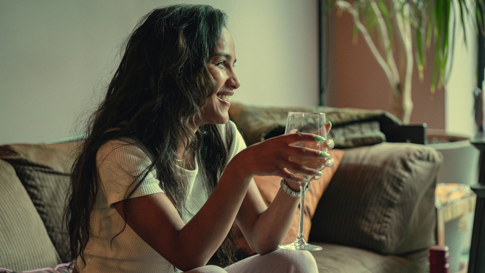 Saffron Hocking as Lauryn in "Top Boy", smiling and sitting on a couch with a glass of wine.