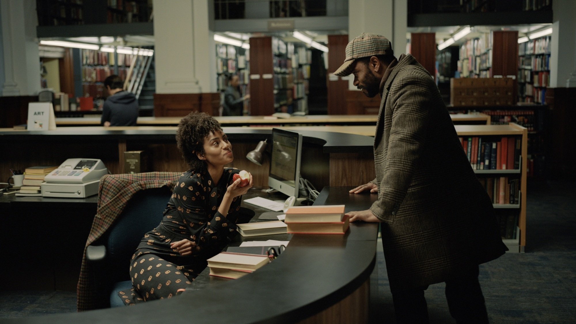 A man leans over a library desk, speaking to the woman sitting behind it.
